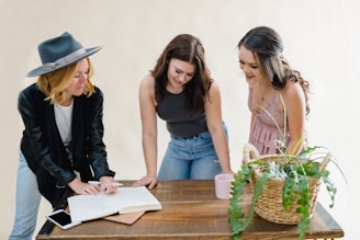 Three women are gathered around a wooden table. One woman is wearing a wide-brimmed hat and a black jacket, writing in a notebook. The other two are leaning over the table, engaged and smiling. A wicker basket with green plants and a pink mug are on the table.