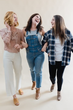 Three women are walking together arm in arm, smiling and laughing. They are dressed in casual attire, with one wearing a pink blouse and white pants, another in denim overalls, and the third in a plaid shirt and black pants. The background is a neutral, light color.