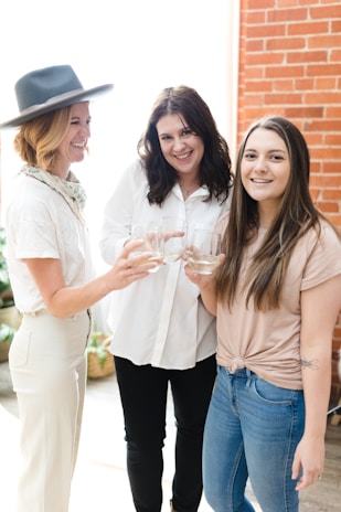 Three women are standing together indoors, smiling and holding glasses as if making a toast. One woman is wearing a hat and light-colored pants, another is in a white shirt, and the third is in a light pink top and jeans. They appear to be in a well-lit room with a brick wall and some greenery in the background.