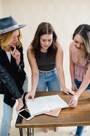 Three women are standing around a wooden table with an open notebook containing handwritten notes. They appear to be engaged in discussion or collaboration. One woman is wearing a gray hat and black jacket, another has a dark sleeveless top, and the third is in a pink sleeveless blouse.