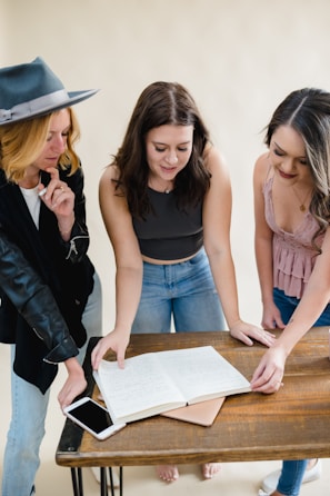 Three women are standing around a wooden table with an open notebook containing handwritten notes. They appear to be engaged in discussion or collaboration. One woman is wearing a gray hat and black jacket, another has a dark sleeveless top, and the third is in a pink sleeveless blouse.