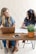 Two women are seated at a wooden table engaged in a conversation. One is using a laptop while the other has a notebook open with a pen in hand. A smartphone is lying on the table, and a potted plant is placed beside them. Both women are smiling, suggesting a friendly atmosphere.