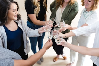 A group of people standing in a circle raising glasses in a toast. They appear to be smiling and enjoying a social gathering. The individuals are casually dressed, and the setting is bright and cheerful.