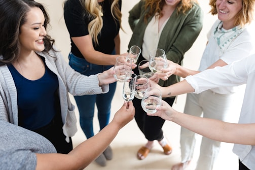 A group of people standing in a circle raising glasses in a toast. They appear to be smiling and enjoying a social gathering. The individuals are casually dressed, and the setting is bright and cheerful.