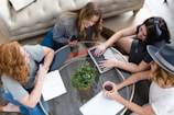A group of four people are gathered around a glass coffee table in a living room setting. One person is using a laptop, another is holding a smartphone, and a third has a notebook. There is a small green plant in the center of the table, and one person is holding a cup. The setting appears informal and collaborative.