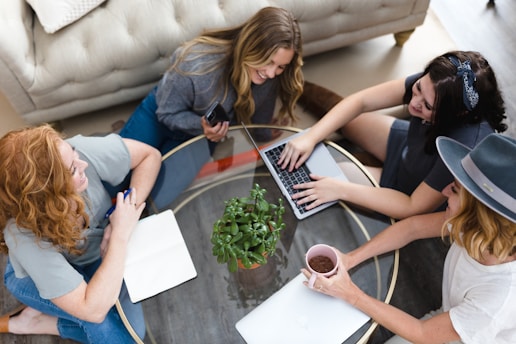 A group of four people are gathered around a glass coffee table in a living room setting. One person is using a laptop, another is holding a smartphone, and a third has a notebook. There is a small green plant in the center of the table, and one person is holding a cup. The setting appears informal and collaborative.