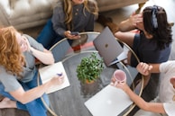 A group of four women are sitting around a circular glass coffee table. One woman is holding a phone, another is writing in a notebook, and a third is gesturing while holding a pink mug. A potted plant is placed at the center of the table, and a laptop is open in front of one of the women.
