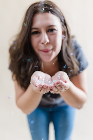 A person with long brown hair is blowing colorful confetti from their cupped hands. The confetti is captured in motion, creating a playful and lively atmosphere. The background is softly blurred, drawing focus on the person and the confetti.