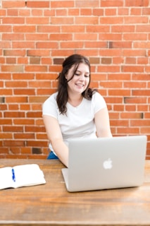 A person wearing a white shirt is seated at a wooden table with a laptop in front of them. An open notebook with a blue pen lies on the table. The background consists of a red brick wall.