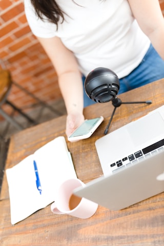 A person wearing a white shirt is using a smartphone with a laptop open on a wooden table. A black microphone is positioned nearby, and there is a notebook with a blue pen and a cup of tea or coffee.