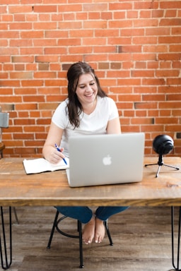 A person writing notes while working on a laptop at a wooden desk
