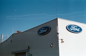 A beige building corner featuring the Ford logo on blue ovals is pictured against a clear blue sky. A person is seen working on the rooftop, surrounded by various rooftop equipment, including antennas and pipes.