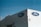 A beige building corner featuring the Ford logo on blue ovals is pictured against a clear blue sky. A person is seen working on the rooftop, surrounded by various rooftop equipment, including antennas and pipes.
