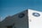 A beige building corner featuring the Ford logo on blue ovals is pictured against a clear blue sky. A person is seen working on the rooftop, surrounded by various rooftop equipment, including antennas and pipes.