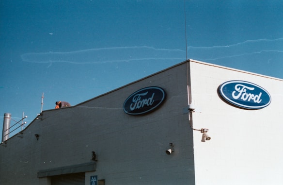 A beige building corner featuring the Ford logo on blue ovals is pictured against a clear blue sky. A person is seen working on the rooftop, surrounded by various rooftop equipment, including antennas and pipes.