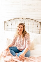 A woman is sitting on a bed with a cozy, comfortable setting. She is casually dressed in a light pink hoodie and blue jeans, smiling warmly. The bed has white and patterned pillows and a light-colored blanket. The background features a whitewashed wooden wall and a metal headboard.