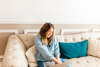 A person with long hair, wearing a denim shirt, is sitting on a beige tufted sofa. They are using a laptop and seem to be smiling, conveying a relaxed atmosphere. The sofa is adorned with a teal pillow and a beige crocheted cushion, set against a beige and white wall with decorative molding.