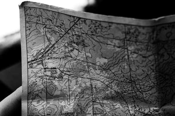 A close-up black and white photograph of a detailed topographic map. The map features contour lines, place names, and various markings indicating geographical features. It appears to be held in a hand, positioned at an angle that highlights its texture and intricate details.