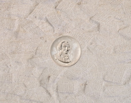 A coin with a profile of a man in a suit, surrounded by text. The background consists of a sandy or textured surface.