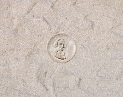 A coin with a profile of a man in a suit, surrounded by text. The background consists of a sandy or textured surface.