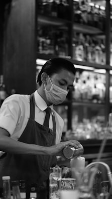 A bartender wearing a face mask is concentrating on preparing a drink. He is dressed in a uniform with a tie and apron, and is surrounded by various bottles and glasses in a dimly lit setting. Shelves filled with bottles line the background.