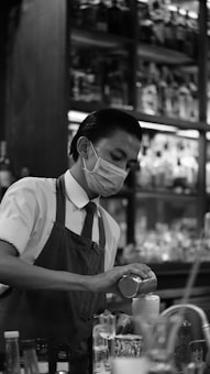 A bartender wearing a face mask is concentrating on preparing a drink. He is dressed in a uniform with a tie and apron, and is surrounded by various bottles and glasses in a dimly lit setting. Shelves filled with bottles line the background.