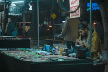A street vendor sits at a stall filled with various colorful handmade jewelry and crafts. The vendor is focused on something in his hands, possibly a phone or small object. Behind the stall, a few people are passing by, and the background is adorned with illuminated signs and lights, creating a dimly lit, urban night atmosphere.