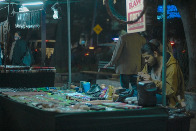 A street vendor sits at a stall filled with various colorful handmade jewelry and crafts. The vendor is focused on something in his hands, possibly a phone or small object. Behind the stall, a few people are passing by, and the background is adorned with illuminated signs and lights, creating a dimly lit, urban night atmosphere.