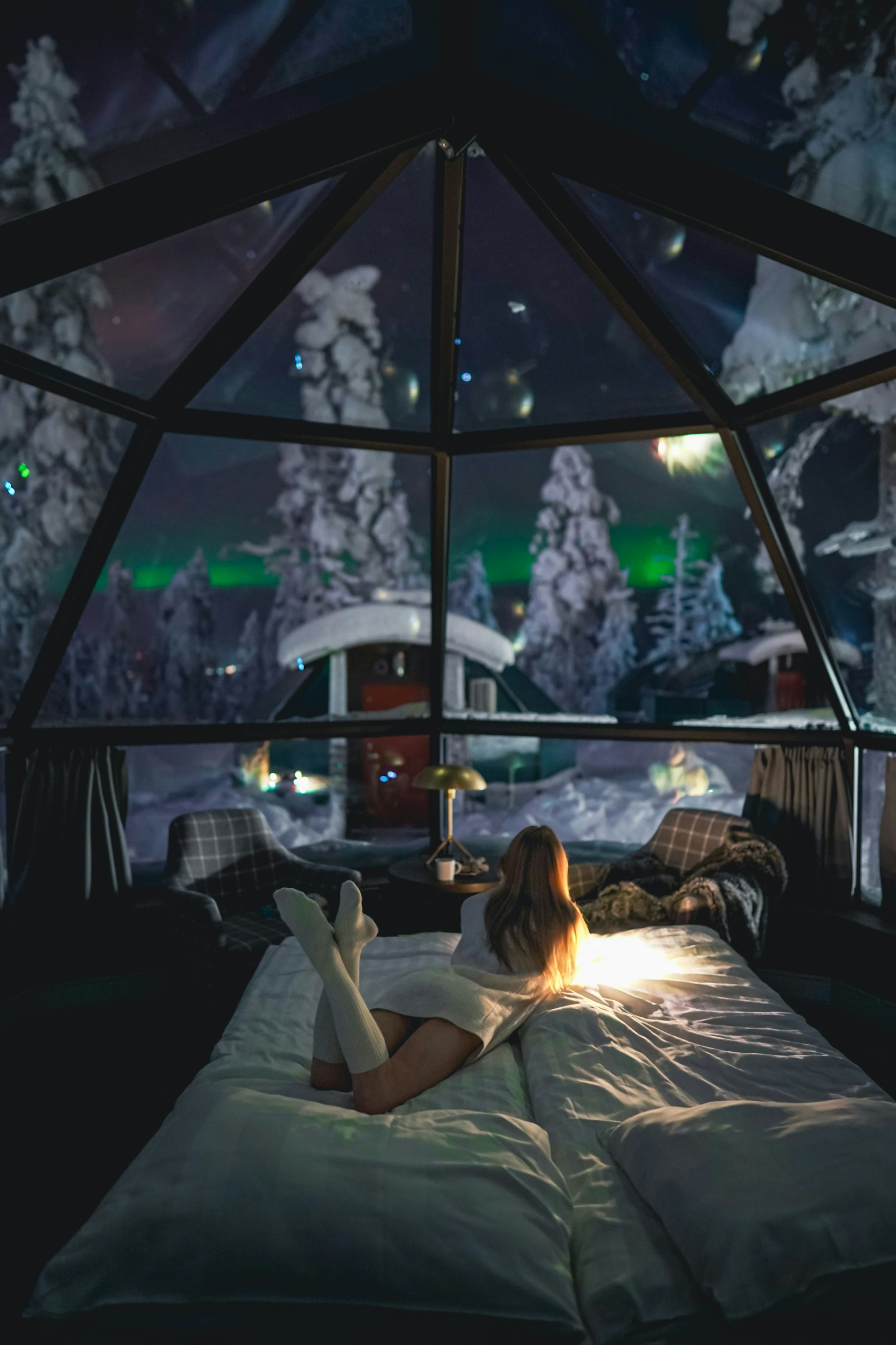 Person inside a glass igloo watching the green aurora borealis through the roof in Finnish Lapland