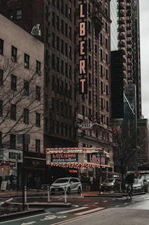 A bustling city street during the day, featuring a prominent theater marquee for 'The Late Show with Stephen Colbert'. The historic buildings display classic architectural details, with a vertical sign on one indicating the word 'COLBERT'. The scene includes cars on the street, street signs, and a cyclist on the road. The atmosphere is moody and urban.