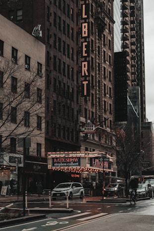 A bustling city street during the day, featuring a prominent theater marquee for 'The Late Show with Stephen Colbert'. The historic buildings display classic architectural details, with a vertical sign on one indicating the word 'COLBERT'. The scene includes cars on the street, street signs, and a cyclist on the road. The atmosphere is moody and urban.