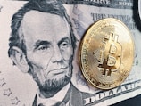 A detailed black and white depiction of a historic figure on a five-dollar bill is prominently displayed. A shiny, gold-colored physical representation of a Bitcoin coin is placed on top of the bill, creating a contrast between traditional currency and digital currency.