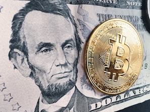 A detailed black and white depiction of a historic figure on a five-dollar bill is prominently displayed. A shiny, gold-colored physical representation of a Bitcoin coin is placed on top of the bill, creating a contrast between traditional currency and digital currency.
