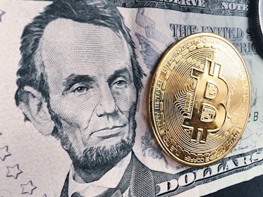 A detailed black and white depiction of a historic figure on a five-dollar bill is prominently displayed. A shiny, gold-colored physical representation of a Bitcoin coin is placed on top of the bill, creating a contrast between traditional currency and digital currency.