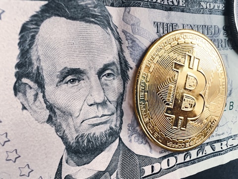 A detailed black and white depiction of a historic figure on a five-dollar bill is prominently displayed. A shiny, gold-colored physical representation of a Bitcoin coin is placed on top of the bill, creating a contrast between traditional currency and digital currency.