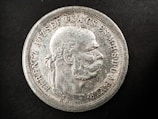 A detailed view of an old coin featuring a profile of a man with a laurel wreath. The coin has raised text along the edges, with intricate detailing around the bust. The surface appears worn and aged with a silver texture against a black background.