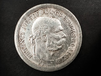 A detailed view of an old coin featuring a profile of a man with a laurel wreath. The coin has raised text along the edges, with intricate detailing around the bust. The surface appears worn and aged with a silver texture against a black background.