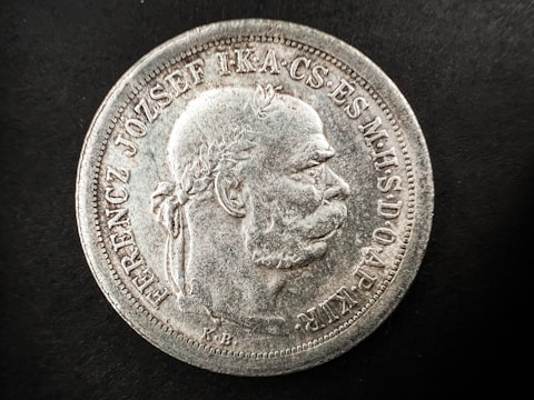 A detailed view of an old coin featuring a profile of a man with a laurel wreath. The coin has raised text along the edges, with intricate detailing around the bust. The surface appears worn and aged with a silver texture against a black background.