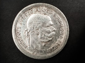 A detailed view of an old coin featuring a profile of a man with a laurel wreath. The coin has raised text along the edges, with intricate detailing around the bust. The surface appears worn and aged with a silver texture against a black background.