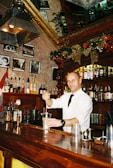 A bartender wearing a white shirt and black tie is mixing a drink behind a wooden bar counter. The bar is decorated for the holidays with garlands and golden bows. Various bottles of alcohol are displayed on the shelves, and several framed black and white photos adorn the wall. The atmosphere is festive and cozy with warm lighting.