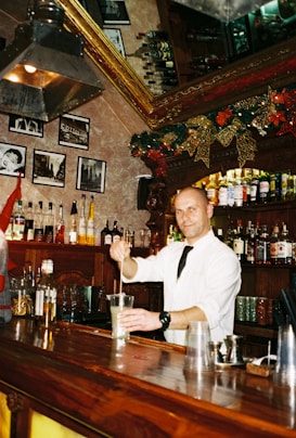 A bartender wearing a white shirt and black tie is mixing a drink behind a wooden bar counter. The bar is decorated for the holidays with garlands and golden bows. Various bottles of alcohol are displayed on the shelves, and several framed black and white photos adorn the wall. The atmosphere is festive and cozy with warm lighting.