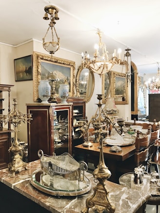 An antique shop interior featuring ornate chandeliers, intricately designed brass and silver items, and elegant wooden furniture. Framed paintings with gold accents are displayed on the walls, and the overall ambiance suggests a vintage, opulent style. The room is filled with a variety of decorative items, including vases and mirrored surfaces, showcasing a rich collection of classic decor.