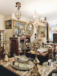 An antique shop interior featuring ornate chandeliers, intricately designed brass and silver items, and elegant wooden furniture. Framed paintings with gold accents are displayed on the walls, and the overall ambiance suggests a vintage, opulent style. The room is filled with a variety of decorative items, including vases and mirrored surfaces, showcasing a rich collection of classic decor.