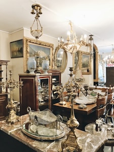 An antique shop interior featuring ornate chandeliers, intricately designed brass and silver items, and elegant wooden furniture. Framed paintings with gold accents are displayed on the walls, and the overall ambiance suggests a vintage, opulent style. The room is filled with a variety of decorative items, including vases and mirrored surfaces, showcasing a rich collection of classic decor.