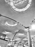The architectural design features a modern ceiling with an intricate pattern of lines and illuminated circular light fixtures. Large columns support the ceiling, and below, there are signs above counters indicating check-in areas or ticketing services.