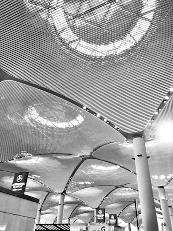 The architectural design features a modern ceiling with an intricate pattern of lines and illuminated circular light fixtures. Large columns support the ceiling, and below, there are signs above counters indicating check-in areas or ticketing services.