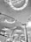 The architectural design features a modern ceiling with an intricate pattern of lines and illuminated circular light fixtures. Large columns support the ceiling, and below, there are signs above counters indicating check-in areas or ticketing services.