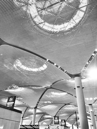 The architectural design features a modern ceiling with an intricate pattern of lines and illuminated circular light fixtures. Large columns support the ceiling, and below, there are signs above counters indicating check-in areas or ticketing services.