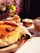 A golden-browned cheesecake rests on a wooden board, surrounded by a setting of fresh flowers in vibrant colors. It is accompanied by a bowl of eggs and another bowl containing a block of butter. The cheesecake has a smooth, creamy texture and is partially sliced, with a decorative serving tool nearby.