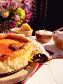 A golden-browned cheesecake rests on a wooden board, surrounded by a setting of fresh flowers in vibrant colors. It is accompanied by a bowl of eggs and another bowl containing a block of butter. The cheesecake has a smooth, creamy texture and is partially sliced, with a decorative serving tool nearby.